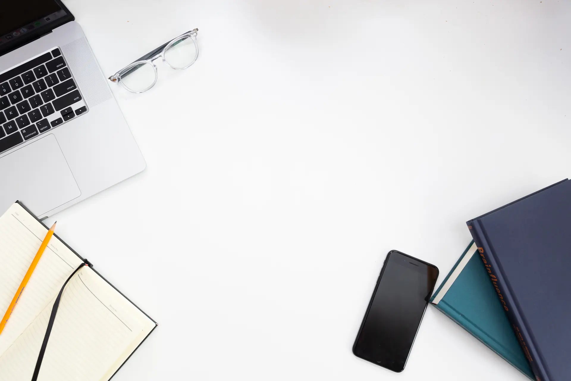 Weiße Schreibtischfläche mit aufgeklapptem Laptop, Brille, Notizbuch mit Stift, Smartphone und zwei übereinanderliegenden Büchern