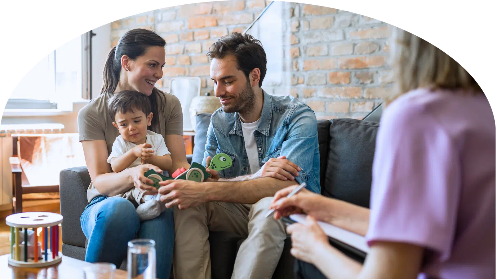 Eltern sitzen mit Kind auf dem Sofa und sprechen mit einer Person, die Notizen macht