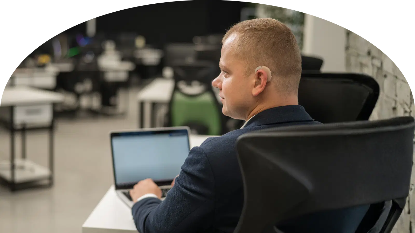 Junger Mann mit kurzem Haar sitzt an einem Tisch und arbeitet an einem Laptop, trägt ein Hörgerät am linken Ohr