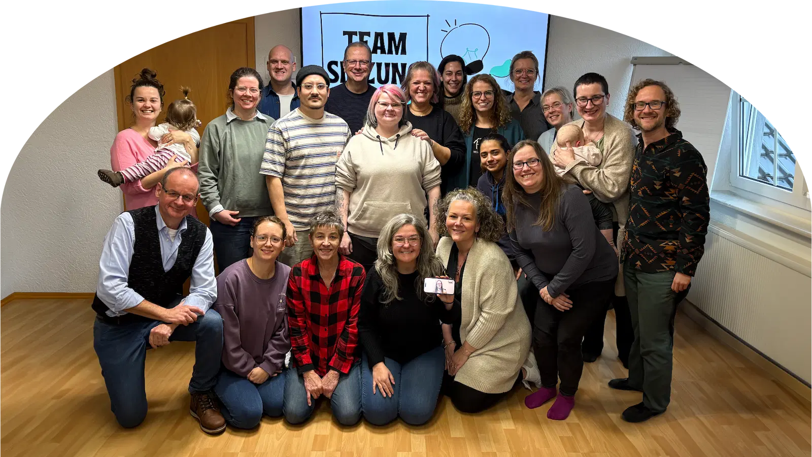 Gruppenfoto des Teams von Hand & Ohr gGmbH mit mehreren Personen in einem Raum vor einer Leinwand mit der Aufschrift 'TEAM'